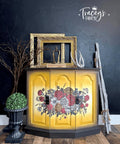 A yellow cabinet with floral designs and black trimmings stands against a dark textured wall. Decorative elements include gilded frames, an antler, twigs, and a potted topiary. The wall features the logo "Tracey's Fancy" with a crown. The wooden floor complements the decor.