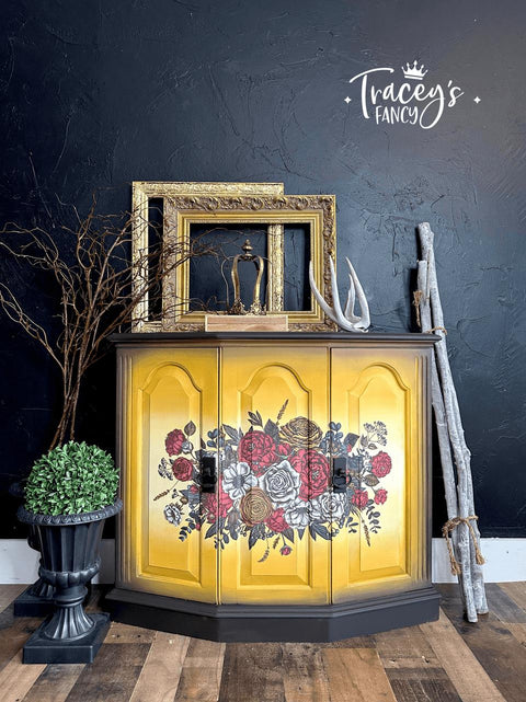 A yellow cabinet with floral designs and black trimmings stands against a dark textured wall. Decorative elements include gilded frames, an antler, twigs, and a potted topiary. The wall features the logo "Tracey's Fancy" with a crown. The wooden floor complements the decor.