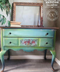 A vintage-style wooden dresser painted in shades of green and adorned with floral designs in the center. An ornate, empty picture frame and a stack of books rest on top. The wall behind shows subtle texture.