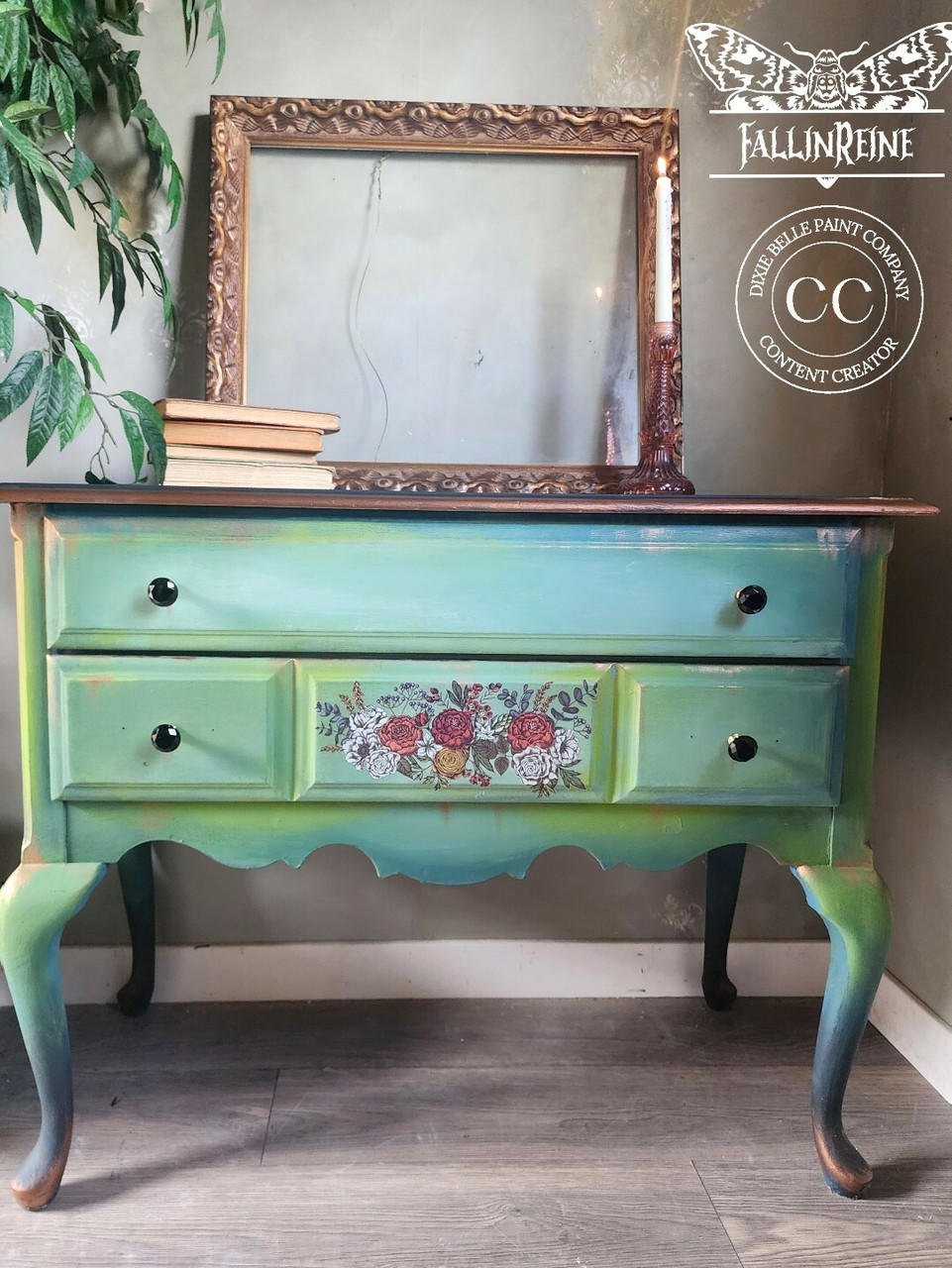 A vintage-style wooden dresser painted in shades of green and adorned with floral designs in the center. An ornate, empty picture frame and a stack of books rest on top. The wall behind shows subtle texture.
