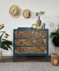 A blue chest of drawers with floral-patterned fronts stands against a white wall. Two hats hang above it. Decorative items such as a vase with a plant, a sculpture, and a candle in a glass holder are on top. Surrounding are potted plants and a wicker lantern on a rug.