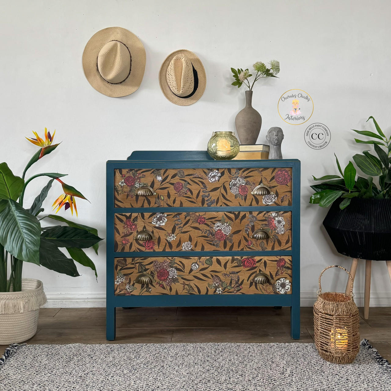 A blue chest of drawers with floral-patterned fronts stands against a white wall. Two hats hang above it. Decorative items such as a vase with a plant, a sculpture, and a candle in a glass holder are on top. Surrounding are potted plants and a wicker lantern on a rug.
