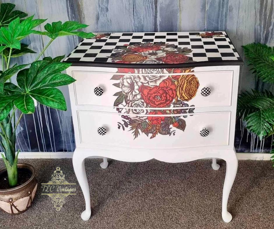 A white ornate dresser stands against a textured gray and blue wall. The dresser features two drawers with floral and checkered patterns. A small potted plant is placed to the left, and part of the wall has cascading paint streaks. The carpet is beige.