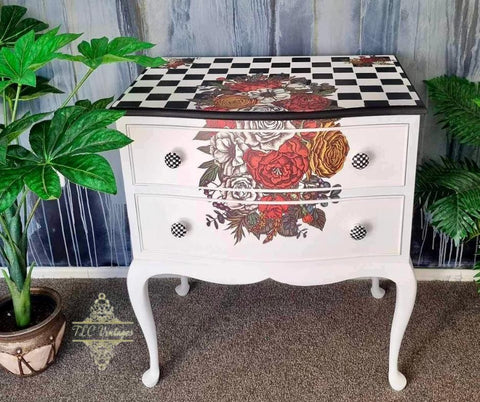 A white ornate dresser stands against a textured gray and blue wall. The dresser features two drawers with floral and checkered patterns. A small potted plant is placed to the left, and part of the wall has cascading paint streaks. The carpet is beige.