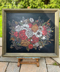 A framed floral design on display shows a bouquet of various flowers, predominantly in shades of red, white, and yellow, with intricate green leaves and stems. The dark background contrasts with the vibrant colors, and sits on an easel outdoors.