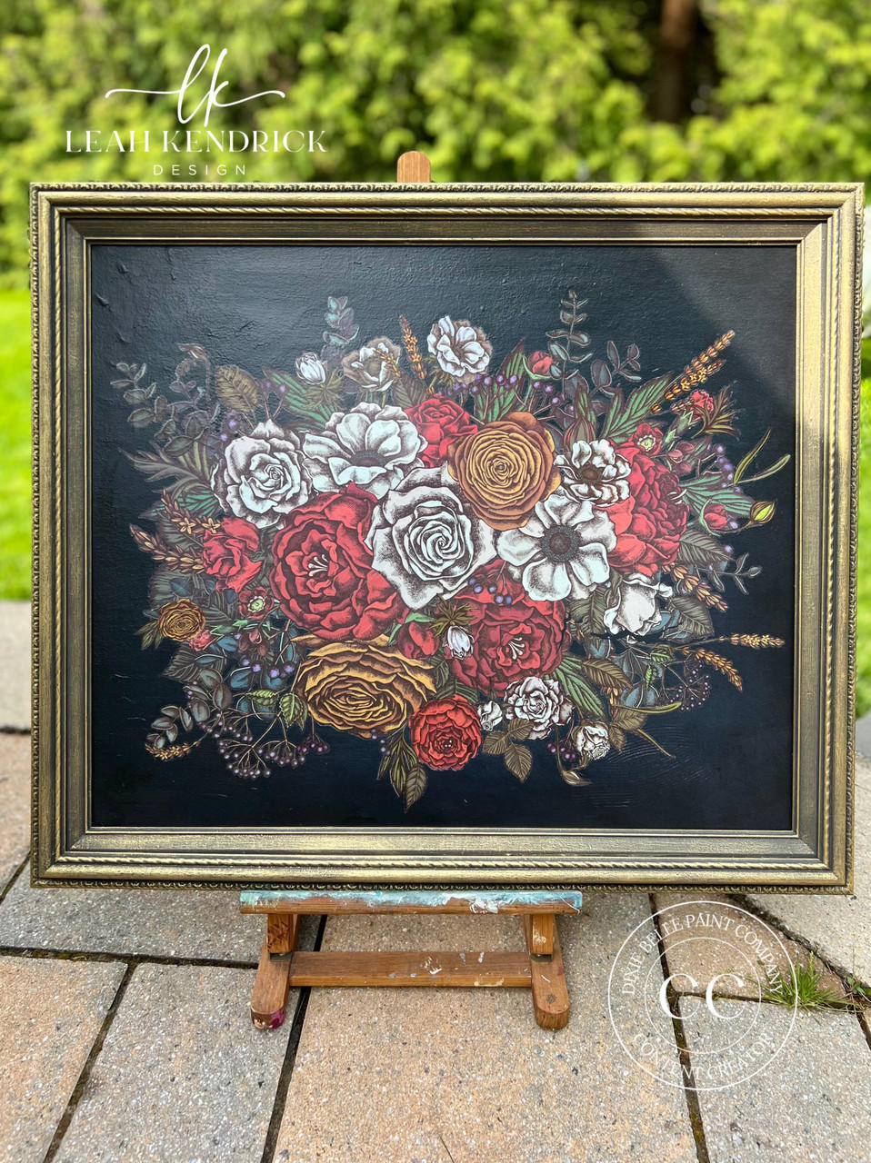 A framed floral design on display shows a bouquet of various flowers, predominantly in shades of red, white, and yellow, with intricate green leaves and stems. The dark background contrasts with the vibrant colors, and sits on an easel outdoors.