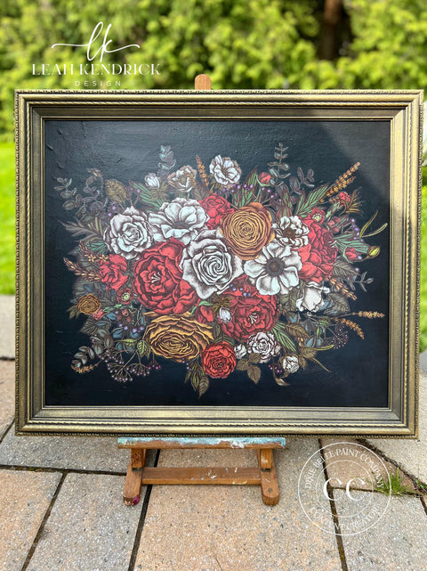A framed floral design on display shows a bouquet of various flowers, predominantly in shades of red, white, and yellow, with intricate green leaves and stems. The dark background contrasts with the vibrant colors, and sits on an easel outdoors.