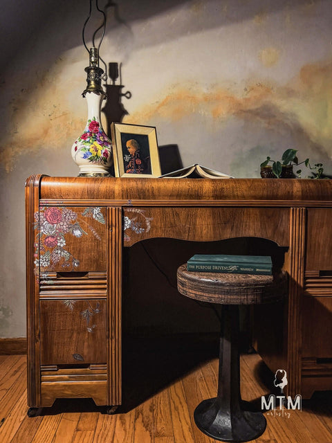 Dark wood desk in front of a faux marble wall, with the Timeless Petals Transfer applied on the drawers. Wooden barstool pulled up to the desk with a book resting on it. Staged with a lamp, picture frame, open book, and pothos plant on top.