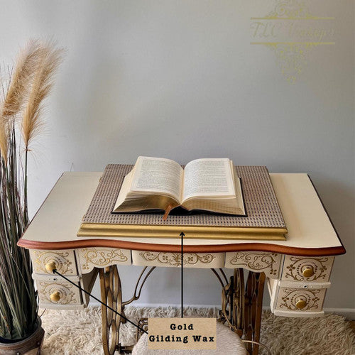 A vintage cream-colored sewing table with ornate gold details holds an open book. Pampas grass stands on the left, and a small label below the table reads "Gold Gilding Wax." A watermark decorates the wall.