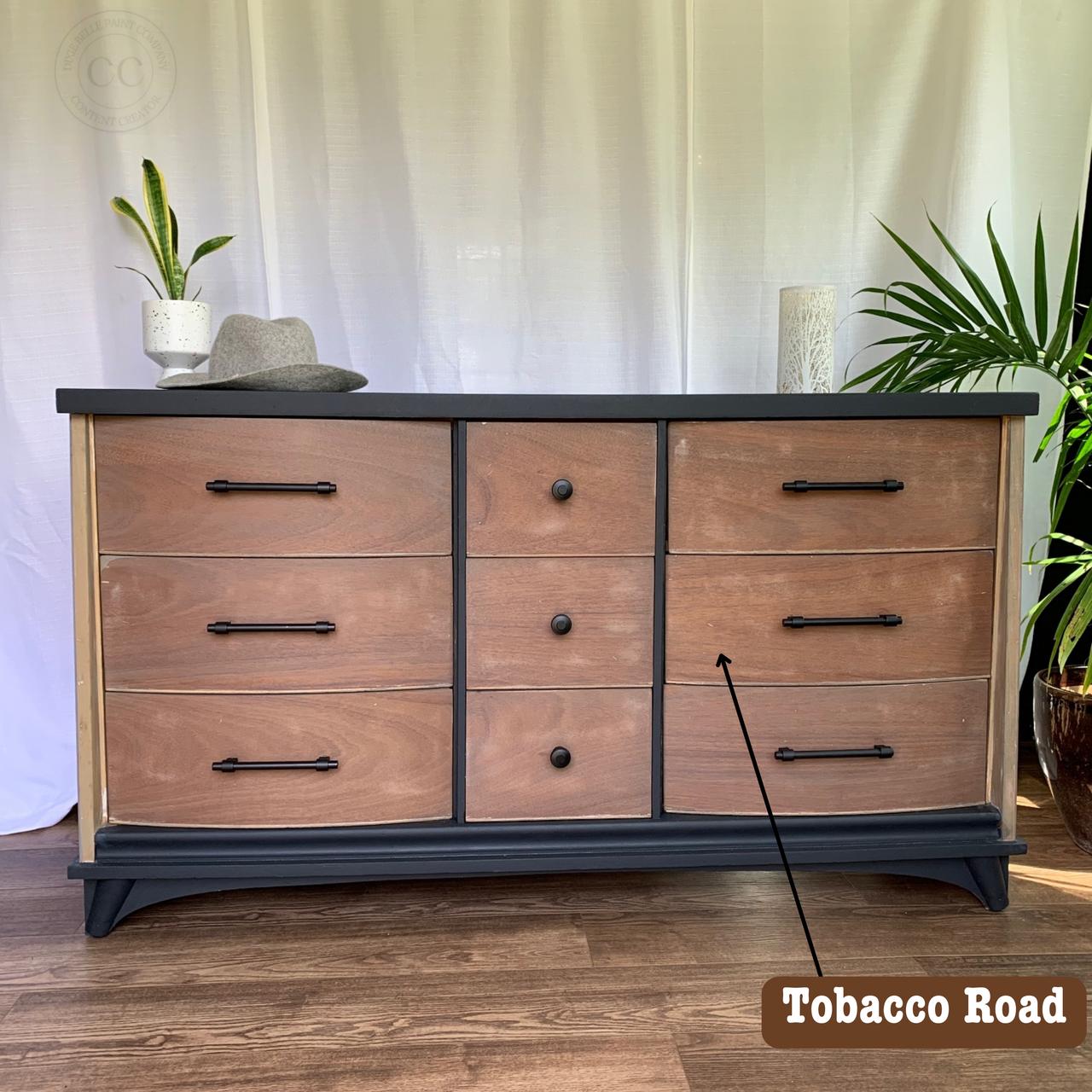 A wooden dresser with nine drawers featuring black handles. A gray hat and snake plant are placed decoratively on top. The words "Tobacco Road" appear over one drawer. A white curtain hangs in the background.