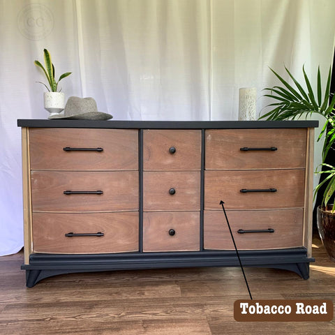 A wooden dresser with nine drawers featuring black handles. A gray hat and snake plant are placed decoratively on top. The words "Tobacco Road" appear over one drawer. A white curtain hangs in the background.