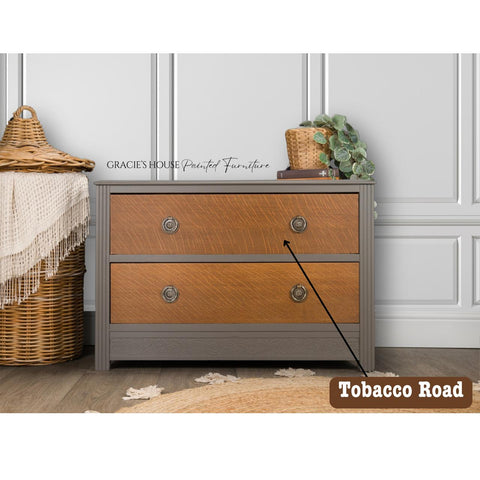 A two-drawer wooden dresser with a gray frame and natural wood front stands against a white paneled wall. A wicker basket with a fringed cloth and a small plant on top add decorative touches. A label reads "Tobacco Road" in the corner.