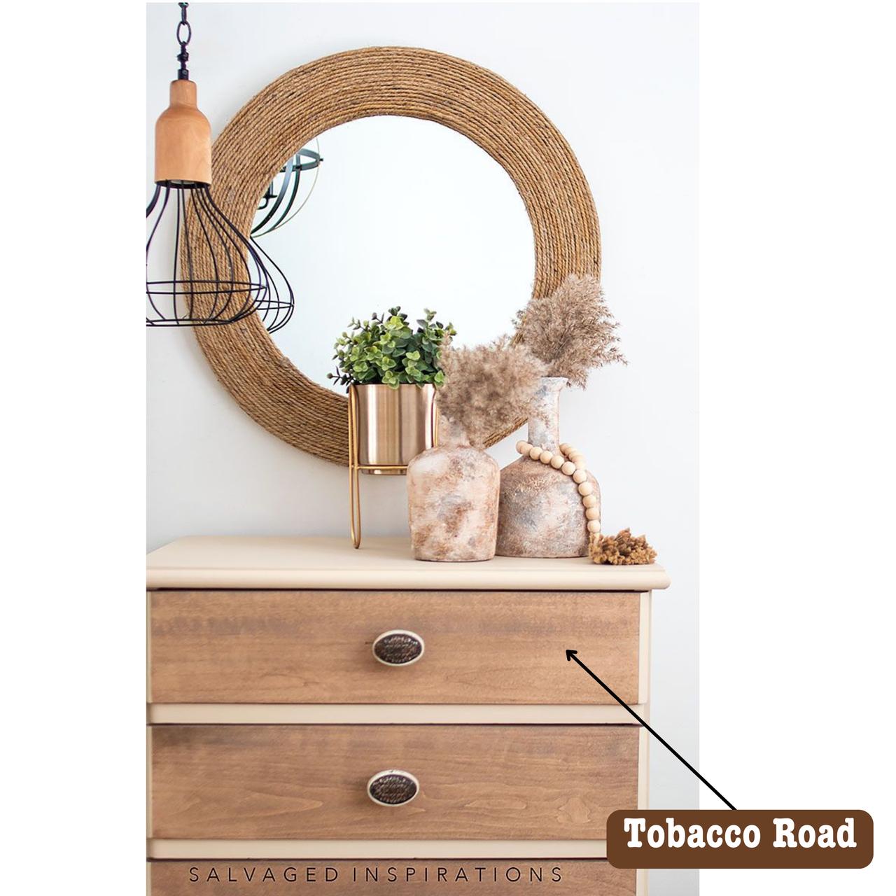 A dresser with a rustic wooden finish, topped with decorative items including a potted green plant, dried pampas grass, and a round mirror with a woven frame. A label reading "Tobacco Road" points to the dresser.
