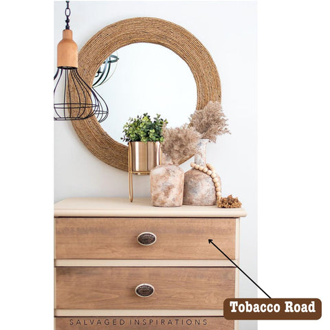 A dresser with a rustic wooden finish, topped with decorative items including a potted green plant, dried pampas grass, and a round mirror with a woven frame. A label reading "Tobacco Road" points to the dresser.