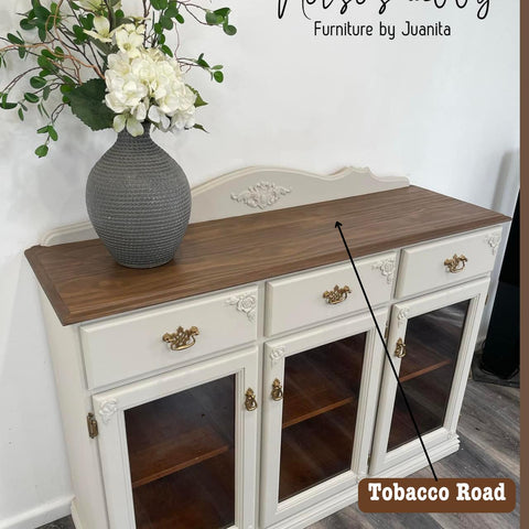 A vintage-style sideboard with a wooden top and white painted cabinets. It has three glass-front cabinets and ornate brass handles. On top, there’s a large decorative vase filled with white flowers and greenery. A label reads "Tobacco Road.