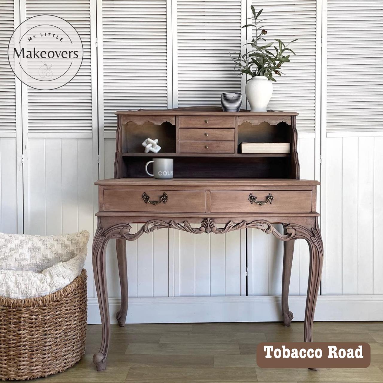 A vintage wooden desk with ornate carvings stands against a white-paneled wall. It features open shelves, small drawers, and a decorative vase on top. A mug is on the desktop. A woven basket is on the floor beside it.