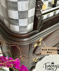 Close-up of an ornate wooden dresser featuring intricate carvings and gold handles. A note points to "Gold Gilding Wax" on the handle. In the foreground, a branch with vivid pink flowers. A black and white checkered pattern is seen on the piece. Logo: "Tracey's Fancy."