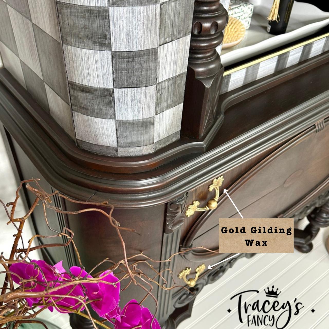 Close-up of an ornate wooden dresser featuring intricate carvings and gold handles. A note points to "Gold Gilding Wax" on the handle. In the foreground, a branch with vivid pink flowers. A black and white checkered pattern is seen on the piece. Logo: "Tracey's Fancy."