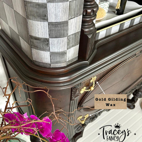 Close-up of an ornate wooden dresser featuring intricate carvings and gold handles. A note points to "Gold Gilding Wax" on the handle. In the foreground, a branch with vivid pink flowers. A black and white checkered pattern is seen on the piece. Logo: "Tracey's Fancy."