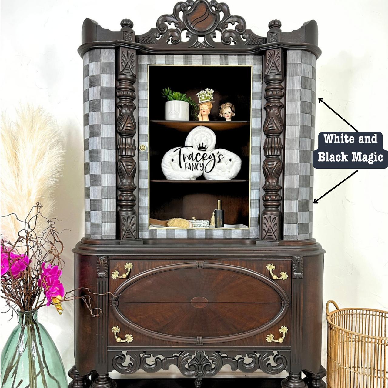 An ornate vintage cabinet with open doors displaying a decorative platter and figurines. The exterior has a checker pattern and intricate woodwork. A nearby vase holds decorative branches and pink flowers. A label reads "White Magic and Black Magic."
