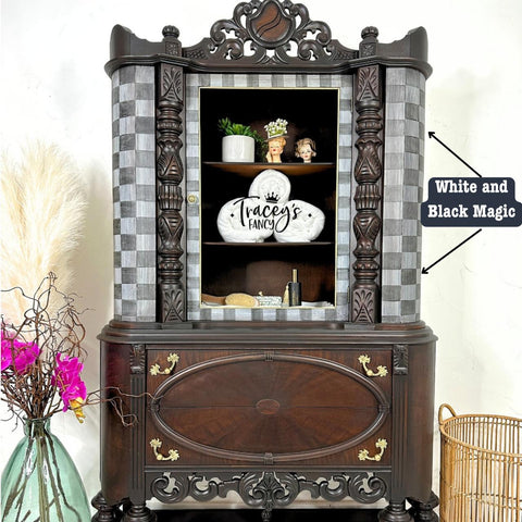 An ornate vintage cabinet with open doors displaying a decorative platter and figurines. The exterior has a checker pattern and intricate woodwork. A nearby vase holds decorative branches and pink flowers. A label reads "White Magic and Black Magic."