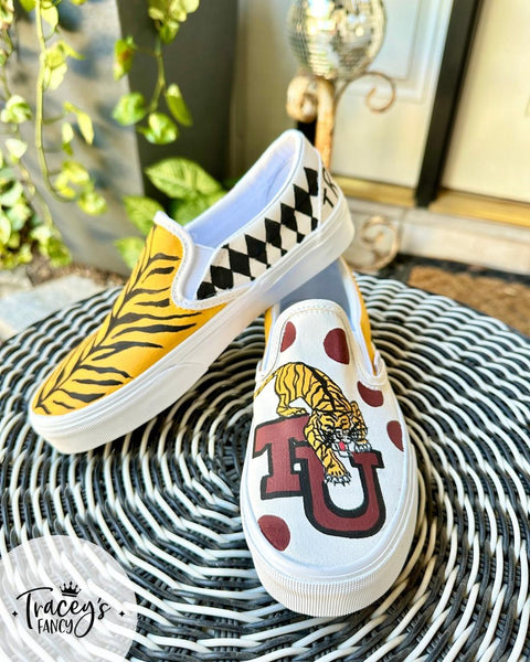 A pair of slip-on shoes on a wicker table. One shoe features a yellow tiger stripe and harlequin design, while the other has a tiger illustration with a red "U" logo and red polka dots. Leafy plant and door in the background.