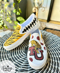 A pair of slip-on shoes on a wicker table. One shoe features a yellow tiger stripe and harlequin design, while the other has a tiger illustration with a red "U" logo and red polka dots. Leafy plant and door in the background.