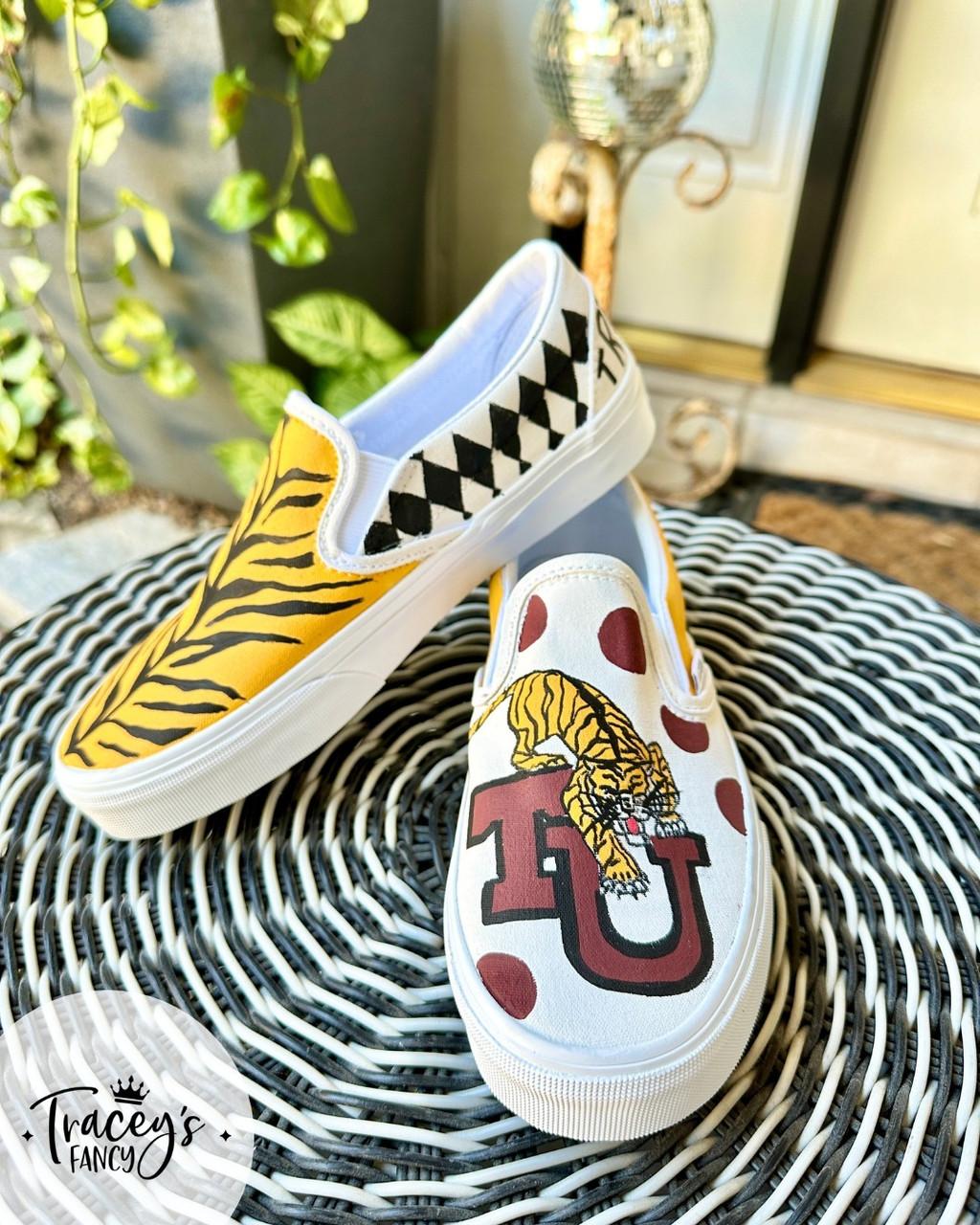 A pair of slip-on shoes on a wicker table. One shoe features a yellow tiger stripe and harlequin design, while the other has a tiger illustration with a red "U" logo and red polka dots. Leafy plant and door in the background.