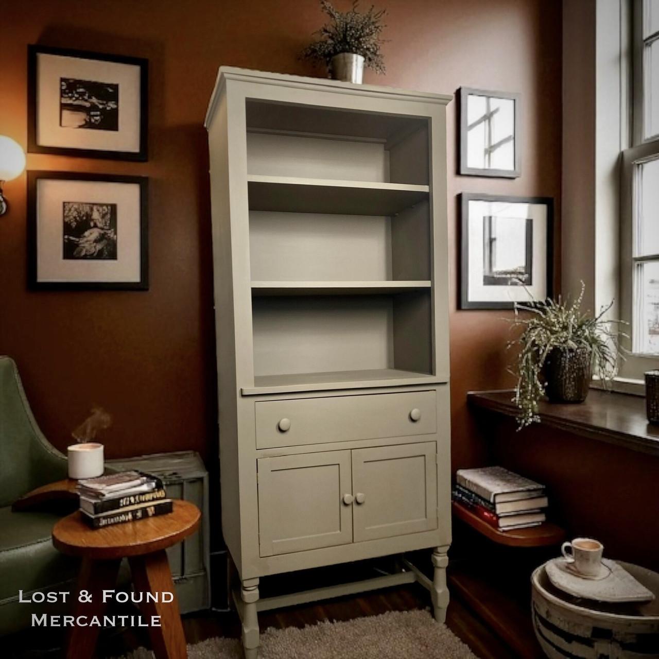 A tall, light brown open-shelf cabinet with two drawers stands in a cozy room with brown walls, framed art, plants, a chair, and a window. Text reads â€œLost &amp; Found Mercantileâ€ in the lower left corner.