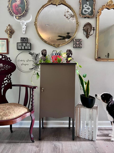A vintage-inspired room with a brown cabinet topped with two busts, a red tin, and plants. Ornate mirrors and framed art hang on the wall. A classic chair sits left, a crate with a plant right, and a black and white dog sniffs nearby.