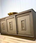 A painted wooden sideboard with three paneled doors, each featuring a rectangular inset design and brass handles, stands on a light wood floor against a white paneled wall. A round brass bowl sits on top.