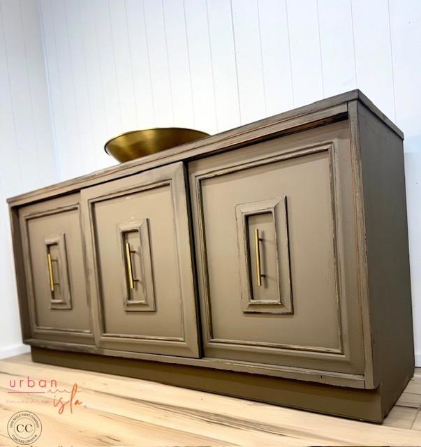 A painted wooden sideboard with three paneled doors, each featuring a rectangular inset design and brass handles, stands on a light wood floor against a white paneled wall. A round brass bowl sits on top.