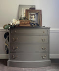 A brown dresser with gold handles stands against a white wall. On top, there are framed pictures, a wooden box, a magnolia flower arrangement, and greenery. The setup is in a softly lit room with wooden floors.