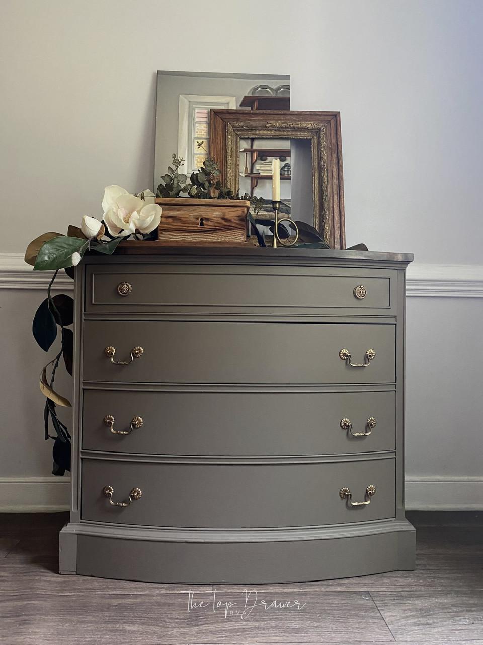 A brown dresser with gold handles stands against a white wall. On top, there are framed pictures, a wooden box, a magnolia flower arrangement, and greenery. The setup is in a softly lit room with wooden floors.
