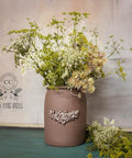 A rustic brown vase with a decorative floral emblem holds an arrangement of delicate white and green wildflowers. Some flowers spill onto the teal surface below. The background is a textured light grey wall with a vintage logo.