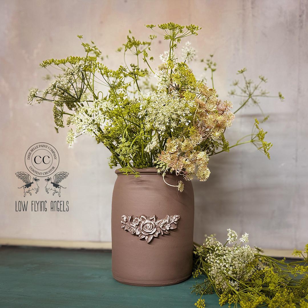 A rustic brown vase with a decorative floral emblem holds an arrangement of delicate white and green wildflowers. Some flowers spill onto the teal surface below. The background is a textured light grey wall with a vintage logo.