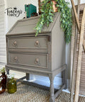 A vintage gray-brown secretary desk with two drawers and ornate drawer pulls sits on a woven rug. A cascading green plant decorates the top, alongside rustic decor and brown glass bottles on the floor.