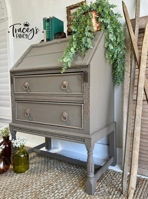 A vintage gray-brown secretary desk with two drawers and ornate drawer pulls sits on a woven rug. A cascading green plant decorates the top, alongside rustic decor and brown glass bottles on the floor.