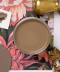 A top-down view of an open can of light brown paint on a floral-patterned surface, with a paint sample card, a paint swatch, and brass decorative objects nearby.