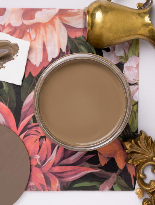 A top-down view of an open can of light brown paint on a floral-patterned surface, with a paint sample card, a paint swatch, and brass decorative objects nearby.