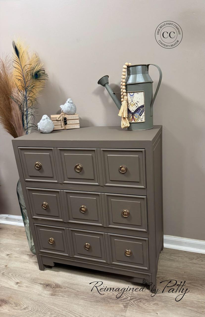 A small gray-brown dresser with gold knobs stands on a wood floor. On top are two ceramic birds, a bundle of books, a watering can with a beaded tassel, and pampas grass in a vase beside it. "Reimagined by Patty" is written in script.