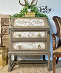 A vintage-style wooden chest with ornate gold details, floral-patterned drawers, greenery, and antlers on top sits against a white wall with "Tracey's Fancy" written on it. A brown chair is partially visible to the right.