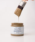 A paintbrush dripping brown paint is held above an open jar labeled "Silk All-In-One Mineral Paint, Truffle." The background is white and minimalistic.