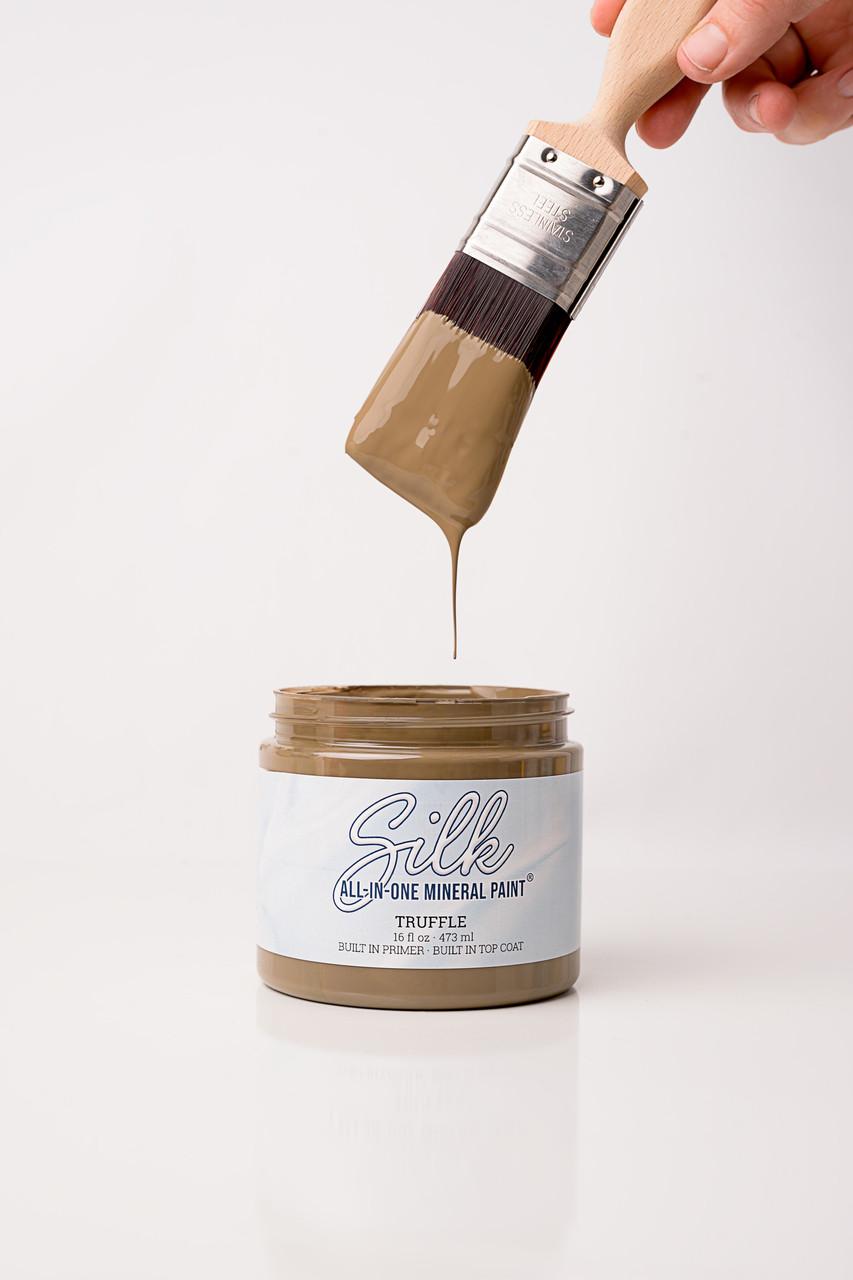 A paintbrush dripping brown paint is held above an open jar labeled "Silk All-In-One Mineral Paint, Truffle." The background is white and minimalistic.