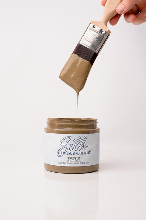 A paintbrush dripping brown paint is held above an open jar labeled "Silk All-In-One Mineral Paint, Truffle." The background is white and minimalistic.