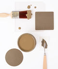 A paintbrush with brown paint, a jar filled with brown paint, a painted square, a round sample, and a palette knife with paint are arranged neatly on a white background.