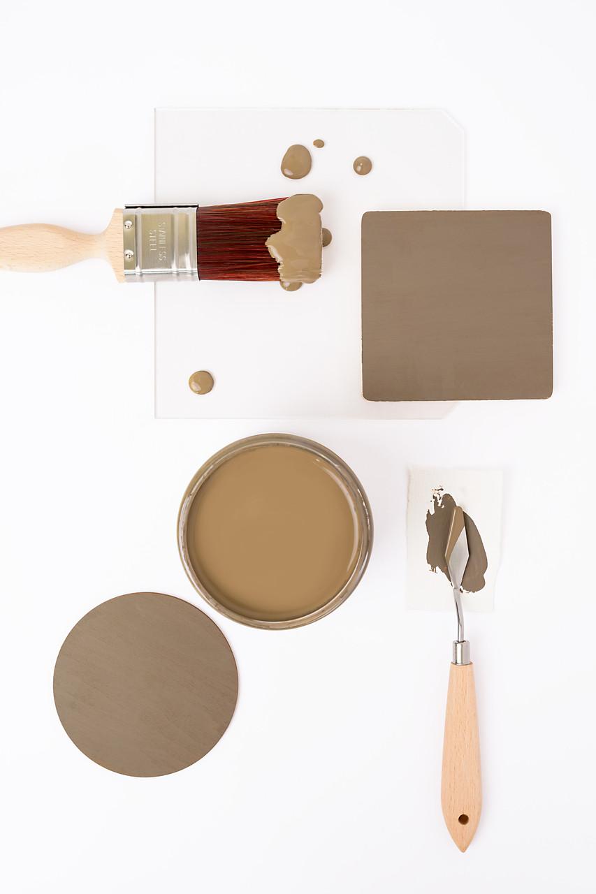 A paintbrush with brown paint, a jar filled with brown paint, a painted square, a round sample, and a palette knife with paint are arranged neatly on a white background.