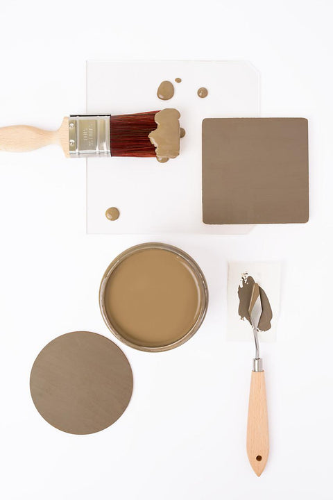 A paintbrush with brown paint, a jar filled with brown paint, a painted square, a round sample, and a palette knife with paint are arranged neatly on a white background.
