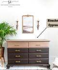 A wooden dresser with six dark drawers and a lighter top in a room with white walls. A potted plant sits to the left, and two wall-mounted candle holders flank a framed painting above the dresser. A label reads "Bleached Cypress.