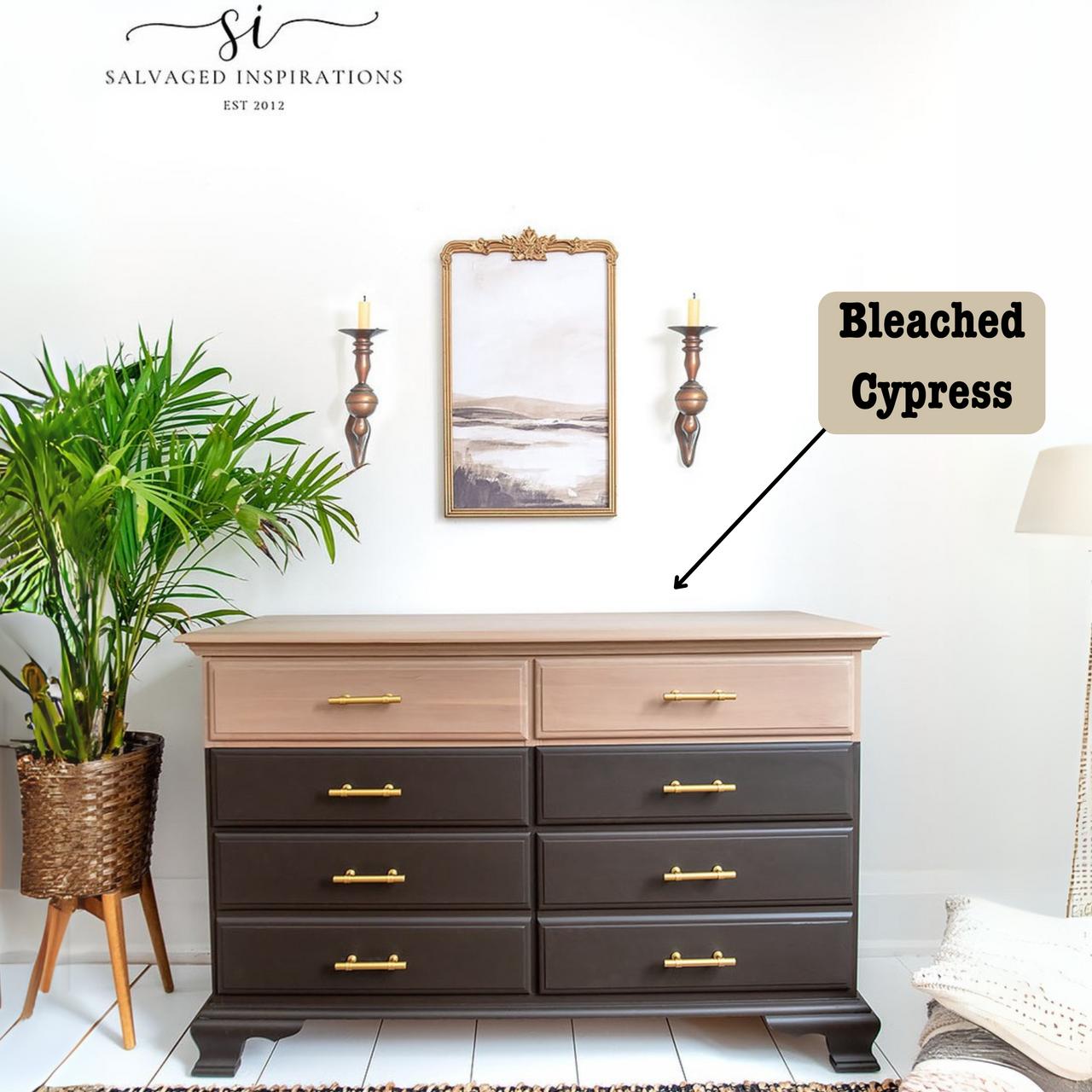 A wooden dresser with six dark drawers and a lighter top in a room with white walls. A potted plant sits to the left, and two wall-mounted candle holders flank a framed painting above the dresser. A label reads "Bleached Cypress.